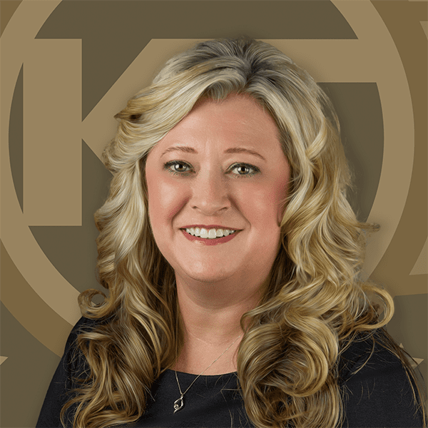 A smiling individual with blonde, curly hair poses in front of a background featuring a large, stylized letter "K".