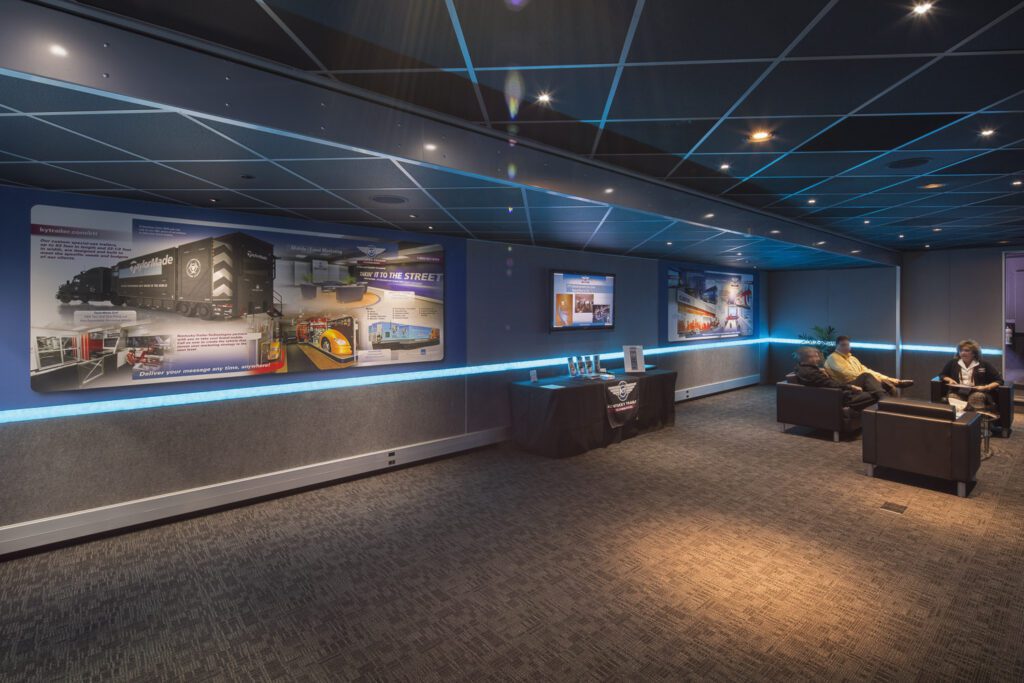 A modern room features neon blue lights crisscrossing the ceiling, shadowy seating areas, sleek dark walls, and various screens displaying content.