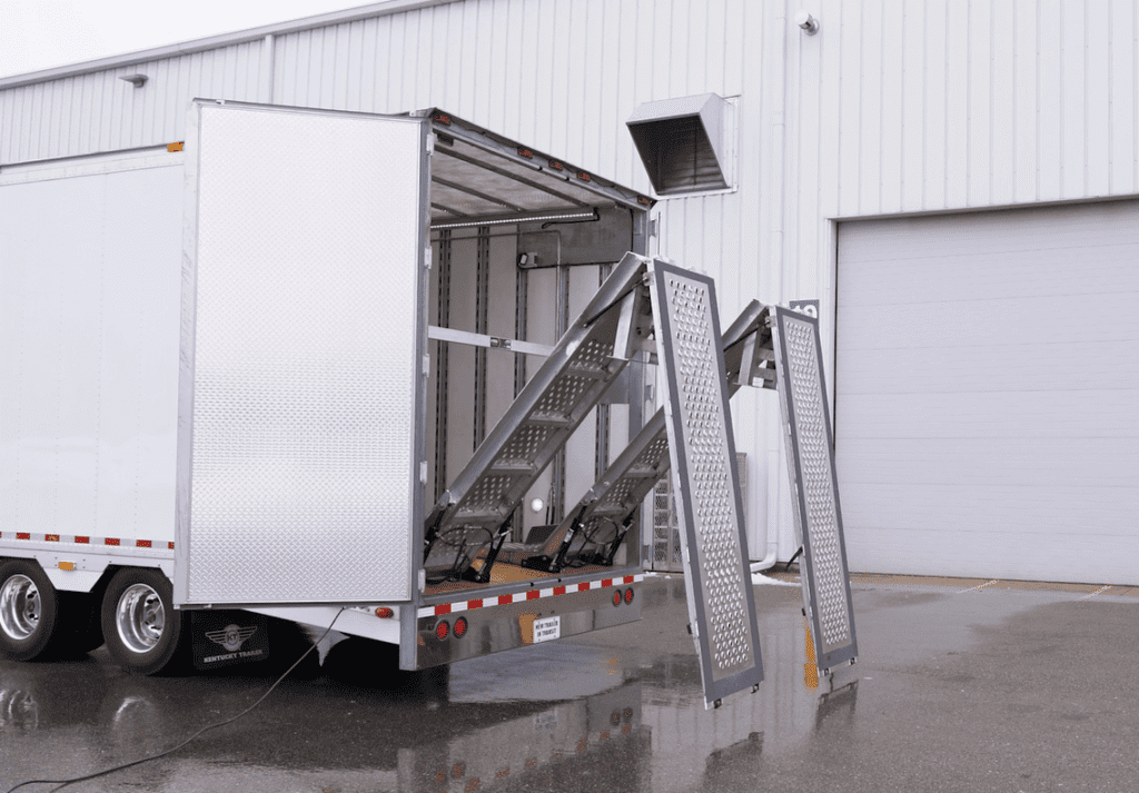 A truck with its rear doors open is parked beside a building, and a metal loading ramp is extended from the truck to the ground.