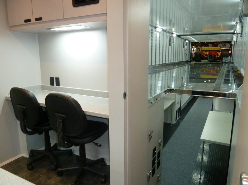 A modern office space features a row of workstations with chairs along one wall and a long, glossy corridor with glass partitions on the opposite side.
