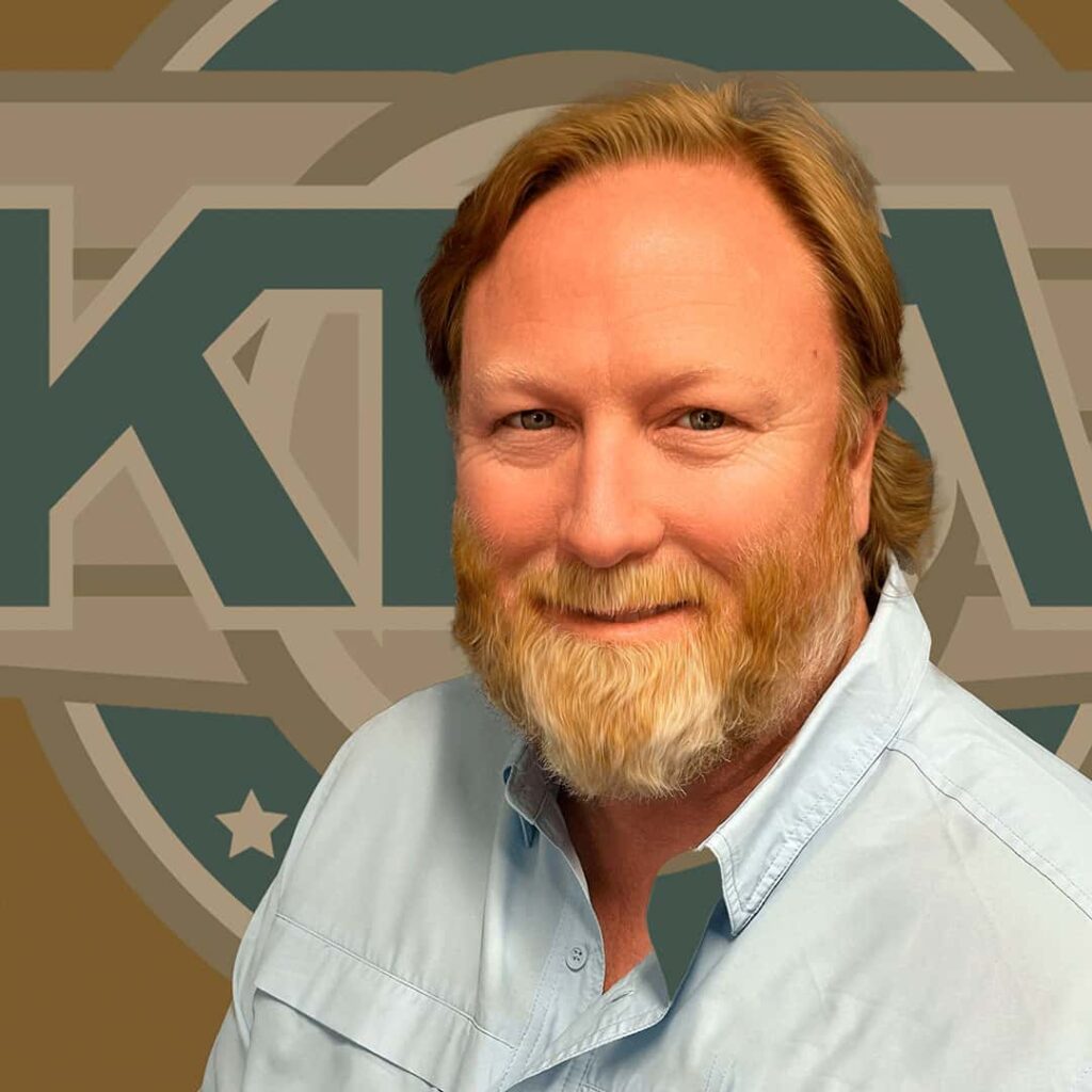A smiling person with a beard and light brown hair is in front of a sign featuring the letters "KTA" and stars.