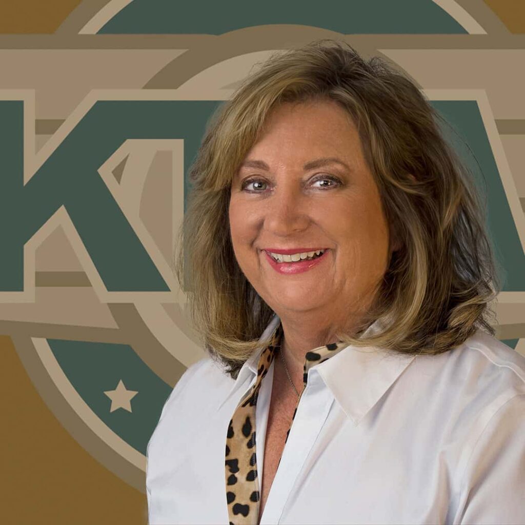 A woman with shoulder-length hair wearing a white blouse smiles in front of a backdrop featuring a large circular logo with the letters "KTV."