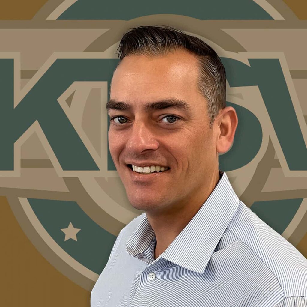 A man with short dark hair and a light blue shirt is smiling in front of a blurred background featuring a circular logo with the word "KAW" visible.