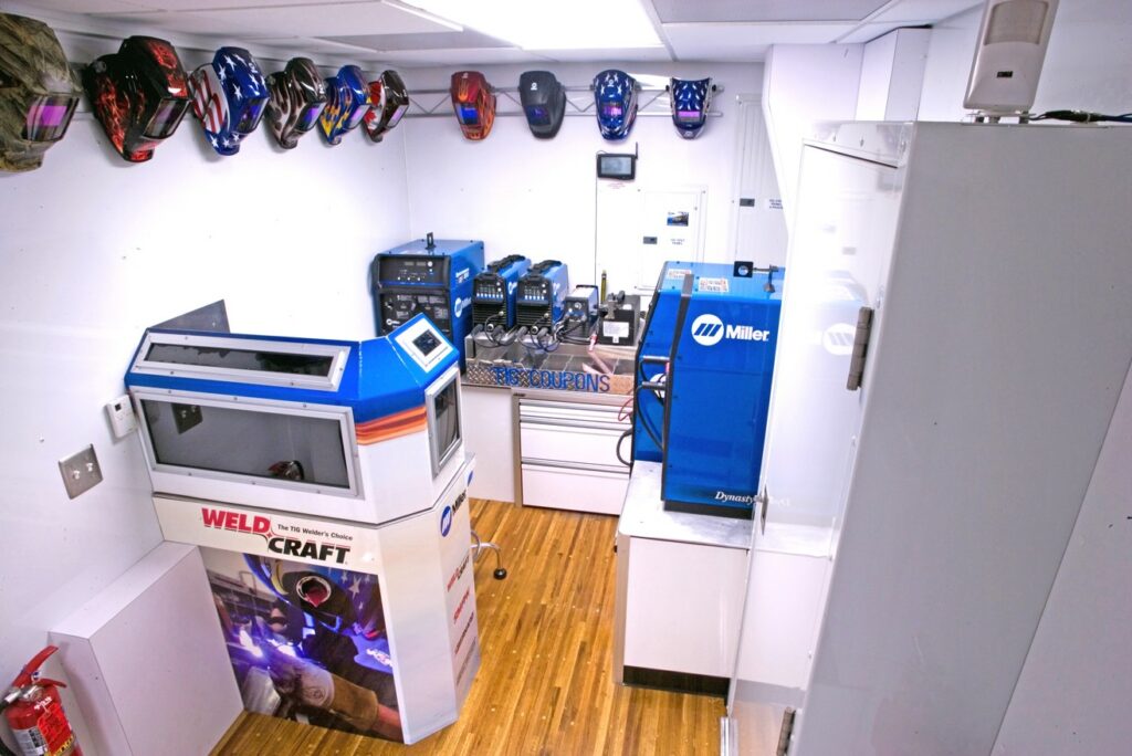 A clean, organized workshop features various machinery, tools, and equipment neatly arranged on shelves and workbenches within a bright, white-walled room with wooden flooring.