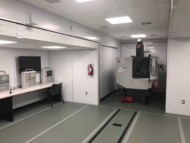 A clean, well-lit laboratory or workshop space features white walls, a long counter with shelves, a fire extinguisher mounted on the wall, and equipment including a large overhead device aligned with tracks on the floor.