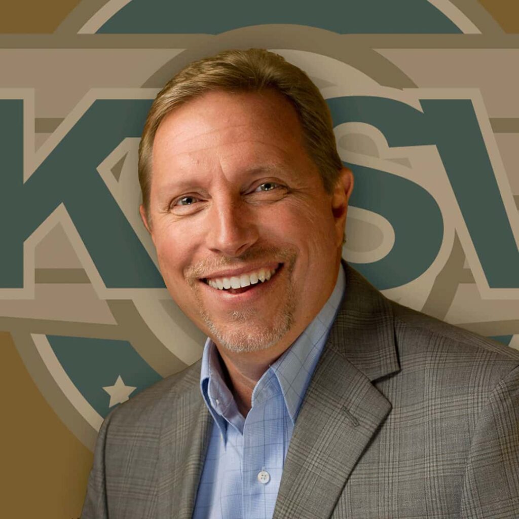 A smiling person wearing a gray blazer and blue shirt is standing in front of a backdrop featuring the letters "KSL."