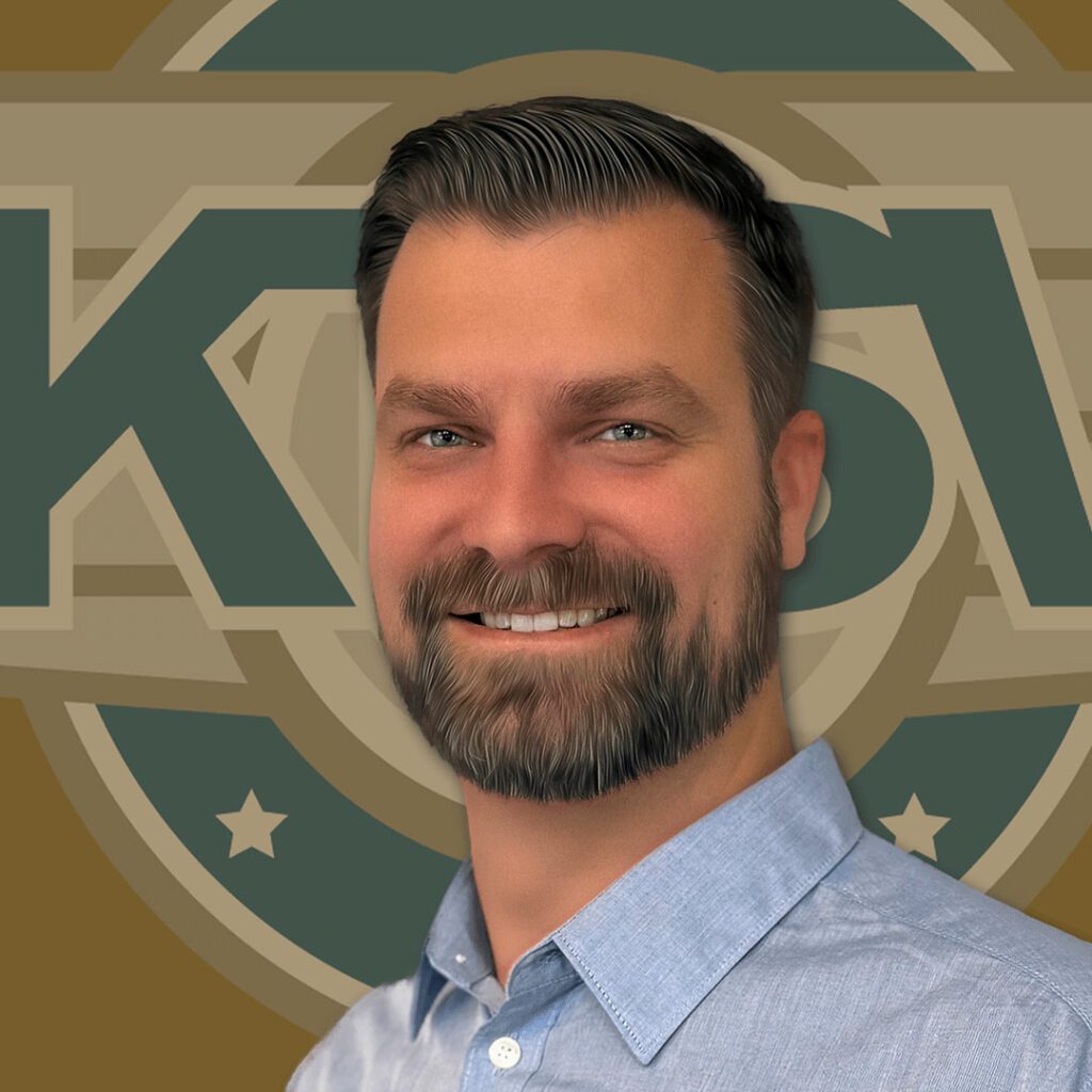 A smiling man with short dark hair and a beard is wearing a light blue collared shirt, standing in front of a logo on the wall.