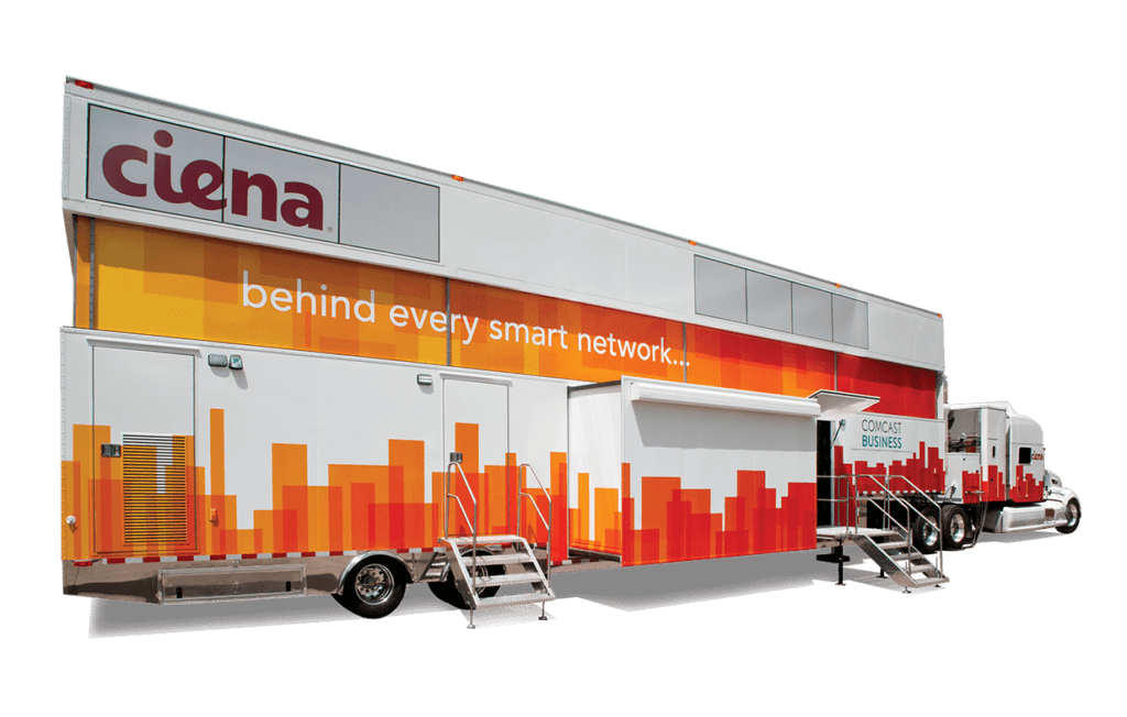 A large mobile unit with red and orange cityscape graphics and the slogan "behind every smart network" is parked outdoors near a building.