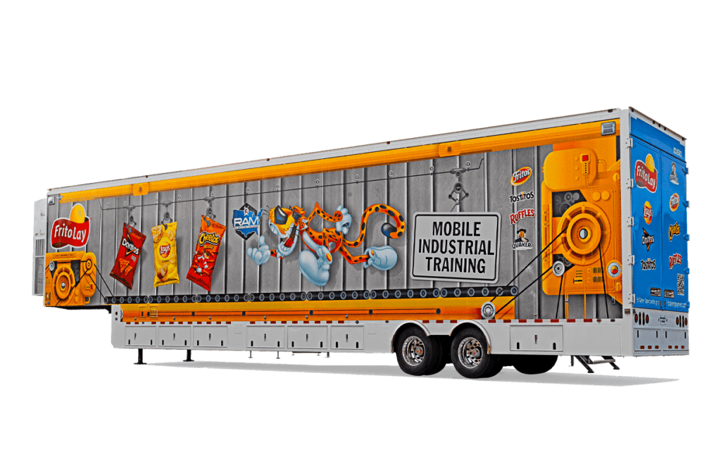 A brightly colored mobile industrial training unit resembling a large trailer is parked outside on a gravel surface.