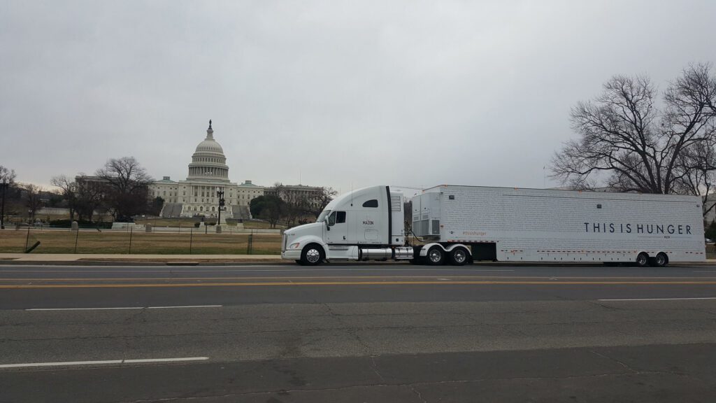 A white semi-truck with a trailer is parked on a street near a large domed government building.